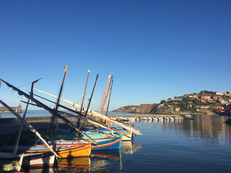Photo of brightly painted traditional wooden fishing boats lined up on a still sea in a harbour.  Clear blue sky overhead.