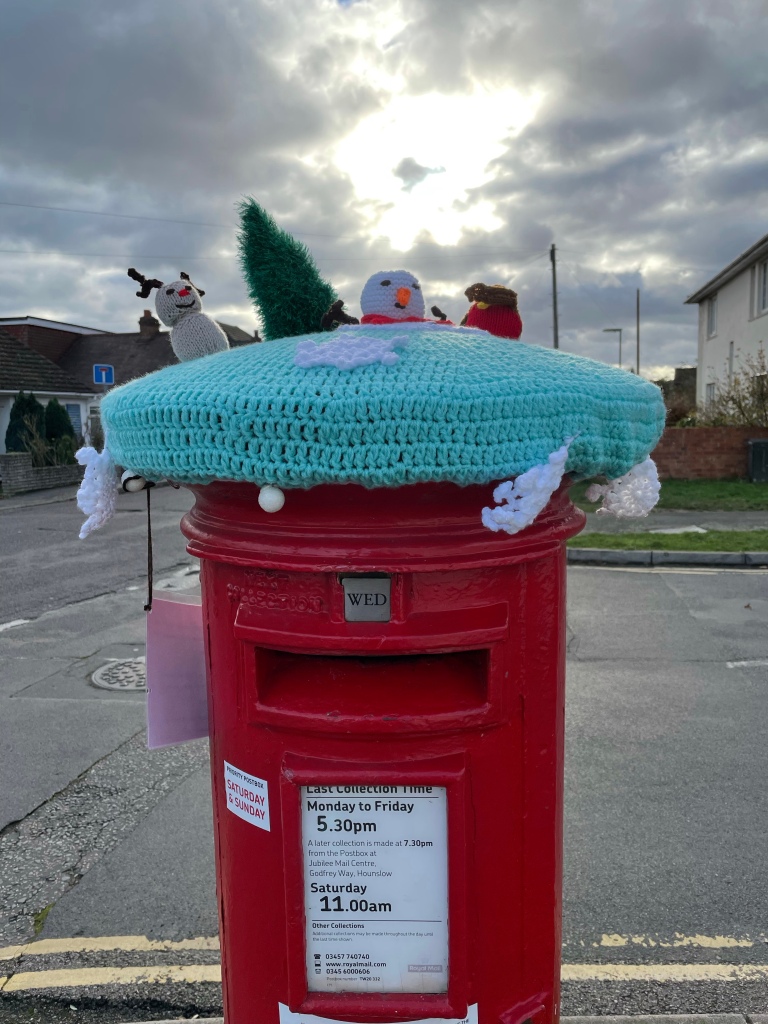 A knitted topper on a circular red post box at a suburban street junction. The topper base is a light turquoise and knitted on top are a reindeer, snowman and robin. Knitted snowflakes hang from the edges.